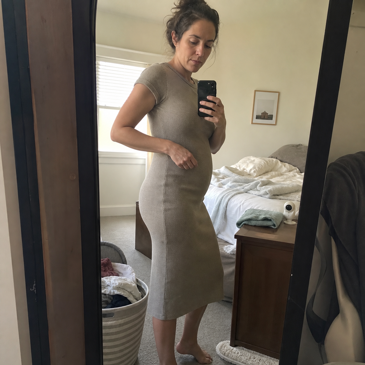 Postpartum mother checking how an everyday knit dress sits in a lived in bedroom